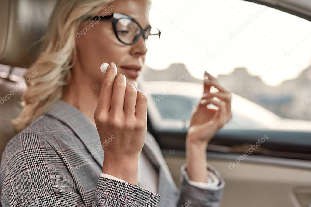 Music is always with me. Attractive and stylish businesswoman in classic wear putting headphones into her ears while sitting on drivers seat in the car