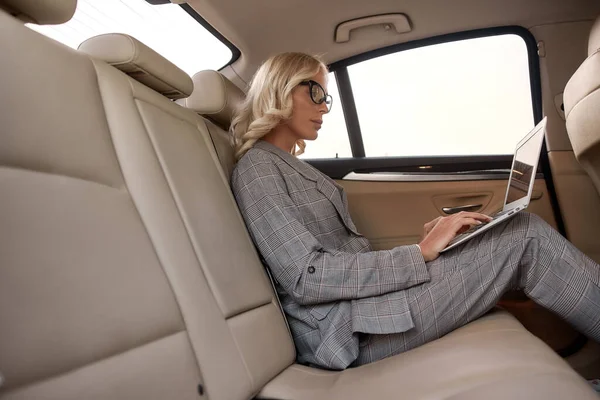 Skupiona bizneswoman. Widok z boku pięknej i pewnej siebie kobiety w pełnym garniturze pracującej na laptopie siedząc w samochodzie — Zdjęcie stockowe