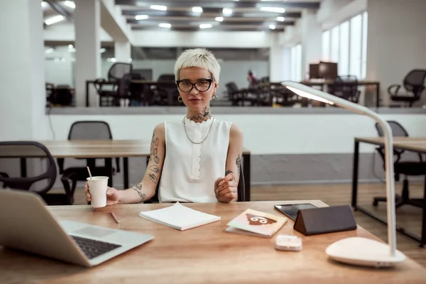 Little break. Young tattooed business woman in eyewear holding cup of coffee and looking at camera while working alone in the modern office