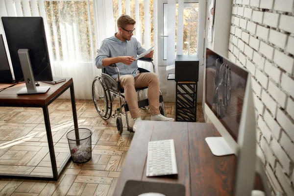 Op zoek naar nieuwe ideeën. Portret van jonge gefocuste mannelijke kantoormedewerker in een rolstoel die een document leest terwijl hij in de buurt van een printer zit op zijn werkplek in het heldere moderne kantoor — Stockfoto