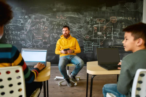 Guiando a la joven generación hacia el éxito. Retrato de un profesor varón escuchando a sus alumnos, mientras estaba sentado con una tableta cerca de la pizarra durante una lección en la escuela inteligente moderna. Enfoque selectivo —  Fotos de Stock