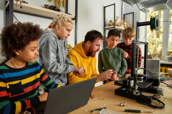 La educación es la herramienta más poderosa. Grupo de niños inteligentes con su profesor masculino mirando la impresora 3d y el prototipo de detalle de plástico en la lección de la escuela de robótica — Foto de Stock