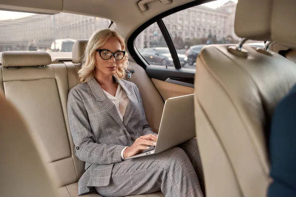 Pracuje gdziekolwiek. Atrakcyjna, skoncentrowana bizneswoman w stylowym klasycznym ubraniu pracująca na laptopie siedząc w samochodzie — Zdjęcie stockowe