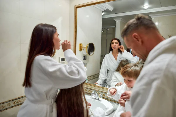 Mas primeiro, higiene. Pais caucasianos e duas crianças de roupão branco tendo rotina matinal no banheiro do hotel, escovando os dentes enquanto estavam perto do espelho. Família, viagens, resort, conceito de férias — Fotografia de Stock