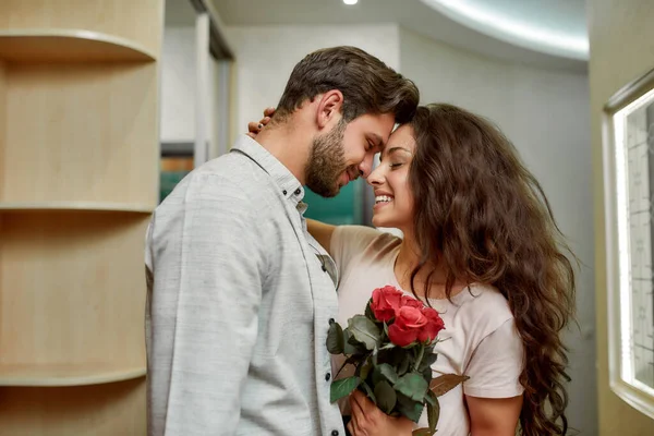 Enamorado. Hombre caucásico guapo trayendo flores a su novia. Feliz pareja abrazándose mientras está de pie en el interior Fotos de stock