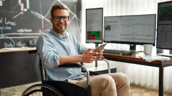 성공 한 무역상. 휠체어에 앉아 스마트폰을 들고 현대 사무실에 앉아 있는 젊고 명랑 한 남성 사업가의 사진. 장애 인 — 스톡 사진