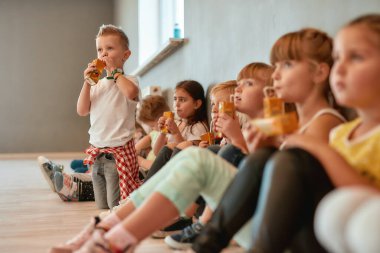 İyileşiyor. Beyaz tişörtlü çocuk dans stüdyosunda dikilirken meyve suyu içiyor. Bir grup tatlı ve pozitif çocuk yerde oturup koreografi dersinden sonra meyve suyu içiyor.