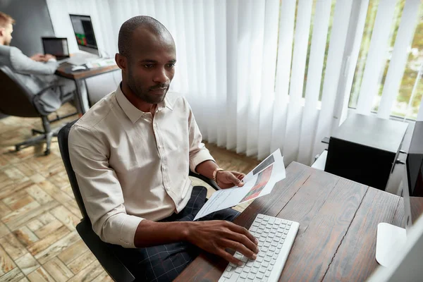 Alles, was Sie brauchen, ist Handel. Blickwinkel auf junge Händler, die am Schreibtisch sitzen und analytische Berichte studieren, während sie im Büro arbeiten. Sein Kollege bedient den Computer im Hintergrund — Stockfoto