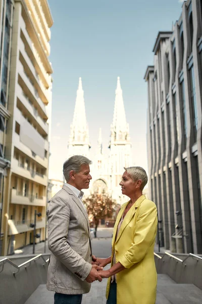 Sempre juntos. Elegante e elegante casal maduro de mãos dadas e olhando um para o outro com sorriso, enquanto em pé contra a bela igreja católica — Fotografia de Stock