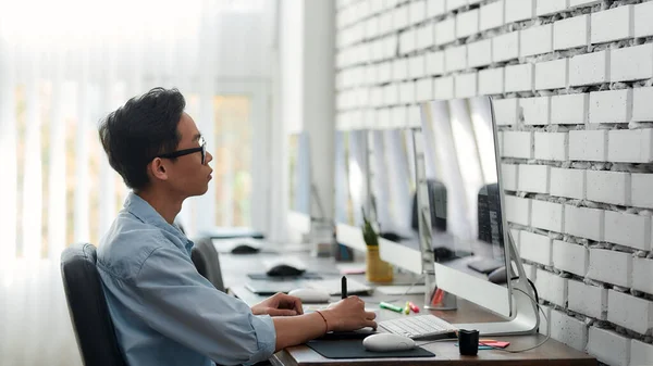 Mantenerse concentrado en el trabajo. Vista lateral de un joven diseñador gráfico asiático en gafas usando una tableta gráfica digital mientras está sentado en su lugar de trabajo en la agencia creativa — Foto de Stock