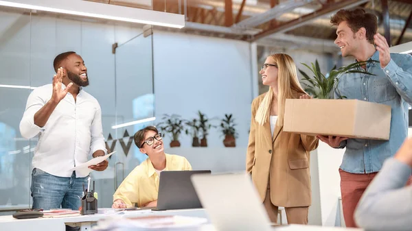Moving to a new office. Young handsome man in casual wear holding box with things and waving to his new coworkers while standing in the modern office — Stock Photo, Image