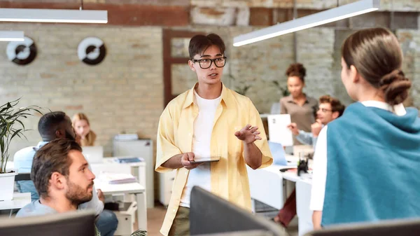 Gran líder de equipo. Joven asiático en ropa casual sosteniendo tableta digital y explicando algo a sus colegas mientras trabajan juntos en la oficina moderna — Foto de Stock