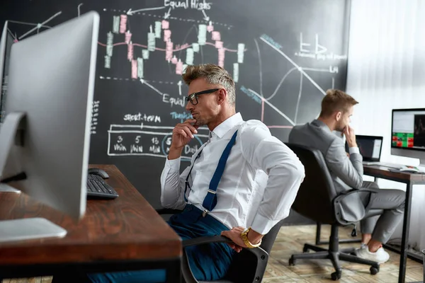 Un gusto per il trading. Trader di mezza età seduto alla scrivania davanti al monitor del computer mentre lavora in ufficio. Il suo collega e lavagna piena di grafici e analisi dei dati in background . — Foto Stock