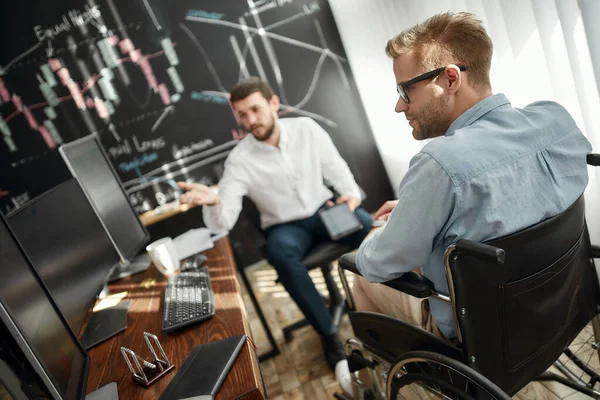 Zakelijke bijeenkomst. Jonge mannelijke kantoormedewerker in een rolstoel bespreekt nieuw project met zijn collega terwijl hij samenwerkt in het moderne kantoor — Stockfoto