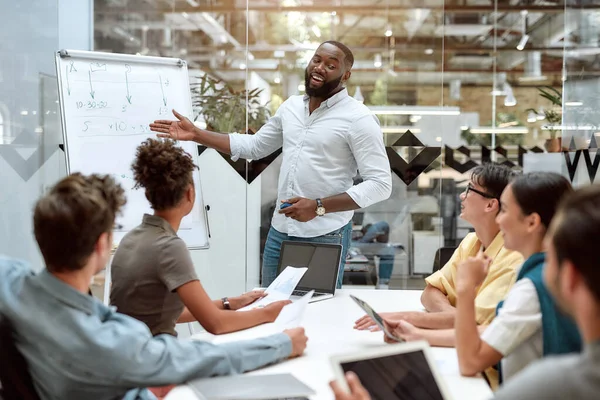 Samen de beste resultaten behalen. Jonge Afro-Amerikaanse zakenman wijst naar wit schoolbord en glimlacht terwijl hij werkt met multicultureel team in het creatieve kantoor — Stockfoto