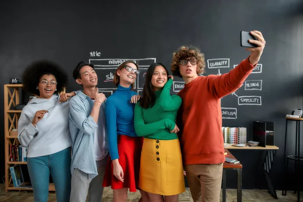 E 'ora dei selfie. Gruppo di persone multiculturali felici in abbigliamento casual fare selfie al telefono mentre in piedi contro lavagna in ufficio moderno. Giovani designer allegri che guardano nella fotocamera — Foto Stock