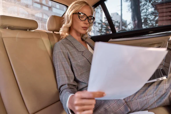 Working from everywhere. Beautiful and successful business woman in full suit analyzing documents and working on laptop while sitting in the car