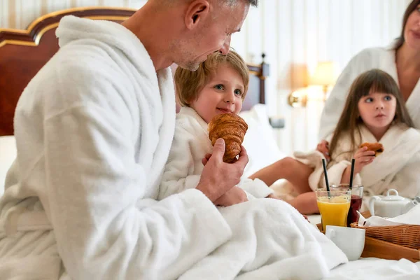 Disfruta el sabor. Primer plano de los padres y dos niños en batas blancas desayunando en la cama, comiendo y bebiendo en la lujosa habitación del hotel. Familia, resort, concepto de servicio de habitaciones — Foto de Stock