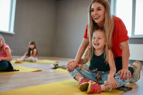 Tränar ett litet barn. Porträtt av en glad kvinnlig tränare hjälpa söt liten flicka att göra fjäril motion. Grupp av barn som sitter på golvet och stretching muskler — Stockfoto