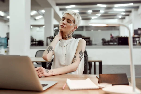 I need a break. Young attractive tattooed business woman feeling tired and massaging her neck while sitting at her working place in the modern office