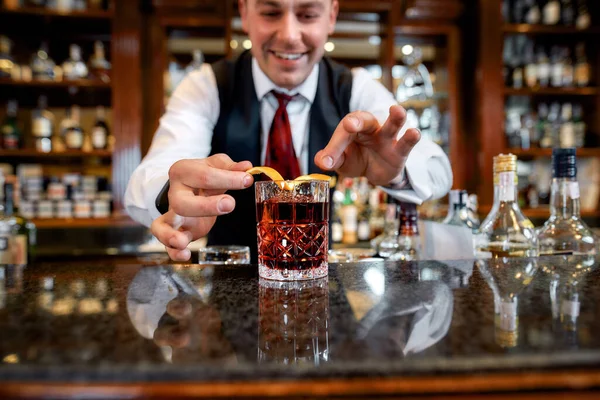 Mantém a calma e a bebida. Jovem barman de pé no balcão, sorrindo enquanto decora coquetel gelado no copo com casca de laranja — Fotografia de Stock
