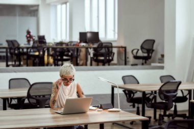 İş yerindeki kadın. Modern ofiste tek başına otururken dizüstü bilgisayarla çalışan, odaklanmış, sarışın, göz dövmeli bir kadın.