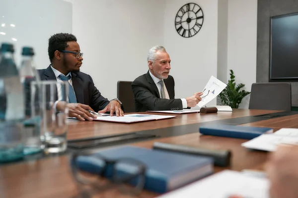 Mira aquí. Hombres de negocios maduros serios analizando documentos y discutiéndolo con colegas mientras tienen una reunión en la oficina moderna — Foto de Stock