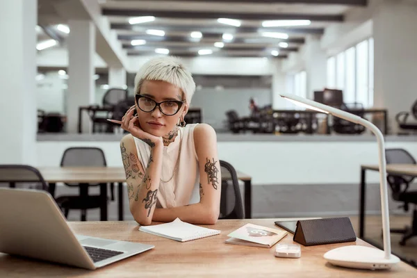 Yaratıcı bir iş. Modern ofiste tek başına çalışırken kameraya bakan güzel ve dövmeli iş kadını.