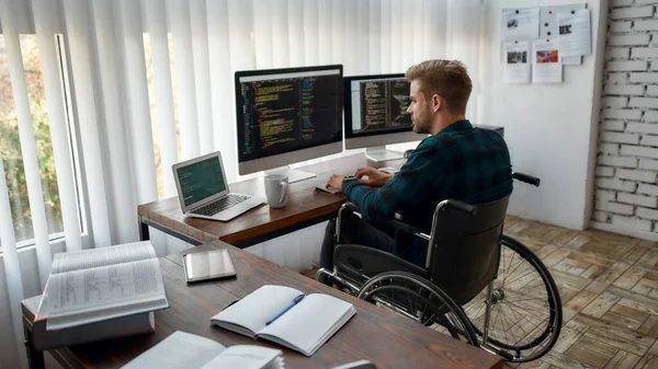 Volledige concentratie. Achteraanzicht van een jonge professionele webontwikkelaar in een rolstoel die programmacode schrijft op meerdere computerschermen terwijl hij op zijn werkplek in het moderne kantoor zit — Stockfoto