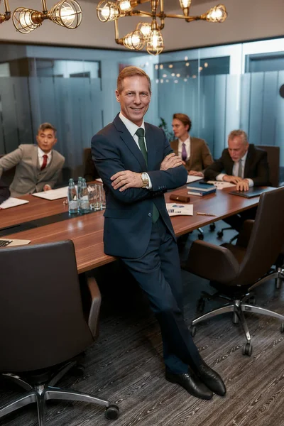 Succesvolle zakenleider. Portret van vrolijke volwassen zakenman in formele kleding houden armen gekruist en kijken naar de camera met glimlach, terwijl leunend op het bureau in het kantoor — Stockfoto
