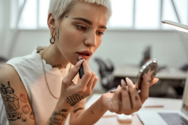 Cute lady at work. Portrait of young and stylish blonde tattooed business woman applying lipstick while sitting in the modern coworking space