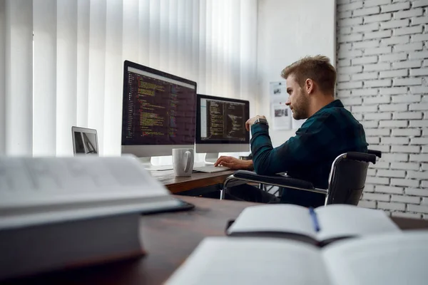 Programador informático en el trabajo. Vista lateral del joven desarrollador web en una silla de ruedas escribiendo código en múltiples pantallas de computadora mientras está sentado en su lugar de trabajo en la oficina moderna. Software de TI — Foto de Stock