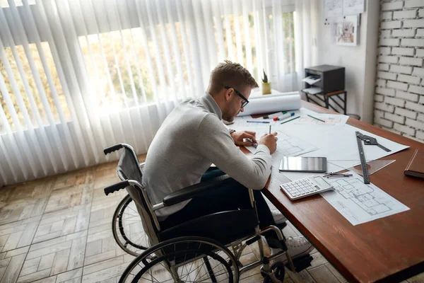 Hard werken. Zijaanzicht van drukke mannelijke ingenieur in een rolstoel die een bouwproject schetst terwijl hij op zijn werkplek in het moderne kantoor werkt — Stockfoto