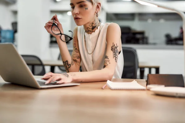 Συγκεντρωμένος στη δουλειά. Νεαρή και όμορφη γυναίκα με τατουάζ δακτυλογραφεί κάτι στο φορητό υπολογιστή, ενώ κάθεται στο χώρο εργασίας της στο σύγχρονο γραφείο — Φωτογραφία Αρχείου