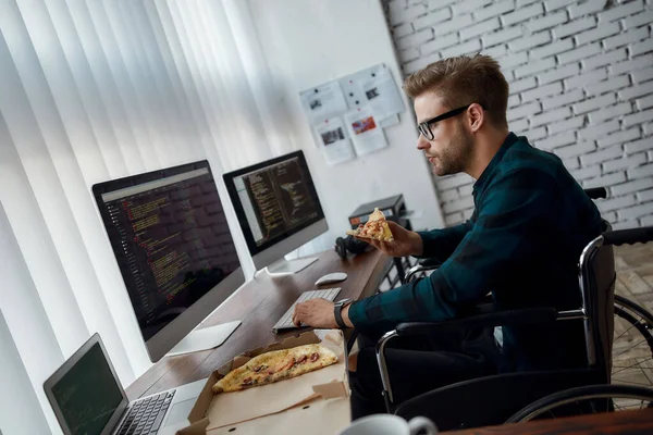 Сосредоточенный мужчина-веб-разработчик в инвалидной коляске ест пиццу и пишет программный код, сидя на рабочем месте с тремя мониторами в современном офисе. Обед — стоковое фото