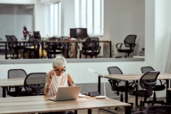 Üzletasszony a munkahelyén. Fiatal szőke tetovált nő szemüveg dolgozik laptop, miközben ül egyedül a modern irodában — Stock Fotó