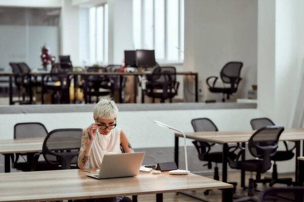 Business lady at work. Young focused blonde tattooed woman in eyewear working with laptop while sitting alone in the modern office