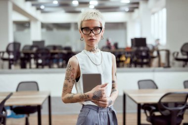 Modern teknolojiler kullanılarak. Gözlüklü, dijital tablet taşıyan ve modern çalışma alanında kameraya bakan genç ve güzel bir iş kadını portresi.