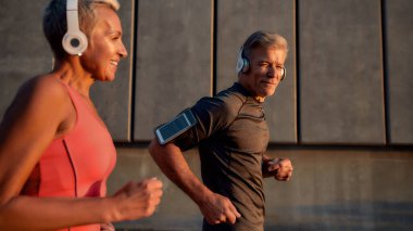 Spor bizi daha mutlu ediyor. Positif ve aktif orta yaşlı kulaklıklı çift sabahın köründe şehir caddesinde koşuşturuyor.