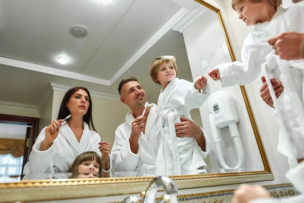 Para sorrisos saudáveis. Pais caucasianos e duas crianças de roupão branco tendo rotina matinal no banheiro do hotel, de pé perto do espelho, indo para escovar os dentes. Conceito de férias em família — Fotografia de Stock