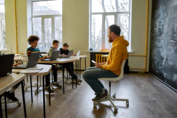 현대의 교훈. 젊은 남성 교사가 자신의 학생들 과 의사소통을 하는 모습, 현대의 똑똑 한 학교에서 수업을 받는 동안 칠판 옆에 태블릿 PC 를 놓고 앉아 있는 모습 — 스톡 사진