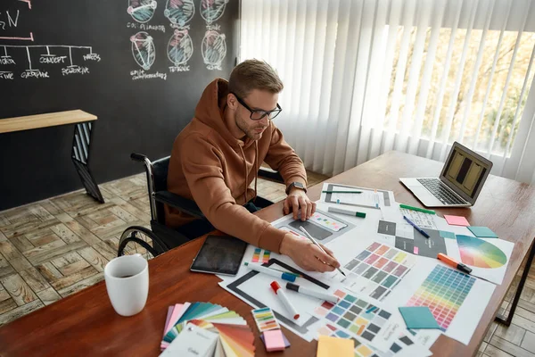 Arbeiten an einem neuen Projekt. Porträt eines professionellen männlichen UX-Designers im Rollstuhl, der an seinem kreativen Arbeitsplatz im modernen Büro die richtigen Farben für die mobile App wählt — Stockfoto