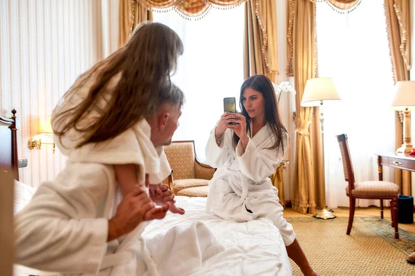 Час зображення. Батьки і дочка в білих халатах проводять ранок разом. Тато і дитина позують на картину, що сидить на ліжку в готельному номері. Сім'я, подорожі, курорт, концепція відпустки — стокове фото