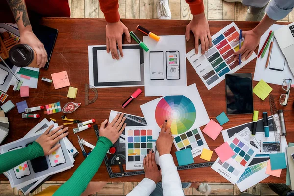 Mobile app design. Top view of designers discussing sketches, choosing colors from palettes lying on the desk while having a meeting in the modern office — Stock Photo, Image