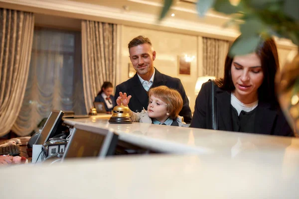 Viajante. Happy family check-in hotel na recepção. Um filho está tocando um sino de serviço enquanto está com os pais na recepção do hotel . — Fotografia de Stock