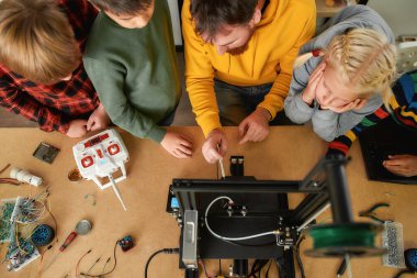 Yaşam için fikirler. Genç erkek öğretmen, robotik okulunda 3D yazıcı olarak çalışan bir grup meraklı çocuğa açıklama yapıyor.