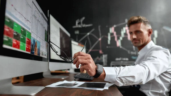 A bright rewarding relationship. Middle-aged trader sitting by desk in front of multiple monitors while working in the office. His colleague and blackboard full of data analyses in background. — Stock Photo, Image