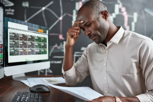 Gana dinero con dedicación. Elegante hombre de negocios africano, comerciante sentado junto al escritorio frente a múltiples monitores. Se está cansando mientras revisa los papeles. . —  Fotos de Stock