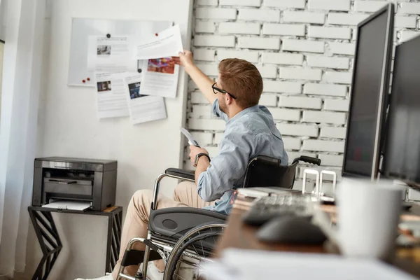 Working day. Young busy male office worker in a wheelchair putting some documents on white board while sitting near printer at his workplace in the bright modern office — Stock Photo, Image