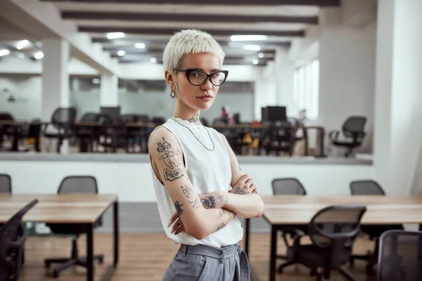 그녀만의 스타일 로. 젊고 매력적 인 금발 문신을 한 사업가가 현대 작업 공간에 서서 팔을 교차하고 카메라를 보고 있는 모습 — 스톡 사진